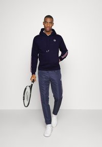 Felpa con cappuccio blu navy con strisce bianche e rosse, abbinata a pantaloni da corsa blu navy con strisce laterali e logo, tenuta con una racchetta da tennis.