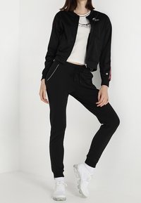 Svart zip-up bomberjacka över en vit T-shirt, kombinerad med svarta joggers. Funktioner inkluderar en figurnära design och vita detaljer. Bärs med vita sneakers.