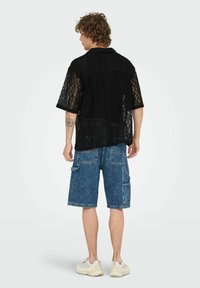 Camisa corta negra de encaje con patrones geométricos, combinada con pantalones cortos cargo de mezclilla azules. El modelo lleva zapatillas ligeras.