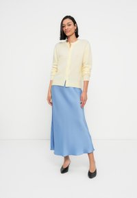 Ανοιχτό κίτρινο ζακετάκι με κουμπιά, φορεμένο πάνω από μια μπλε σατέν φούστα μέση, συνδυασμένο με μαύρα παπούτσια με χαμηλό τακούνι. Λείες υφές και άνετη γραμμή.