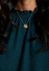 Guldberlockhalsband med intrikat design på en fin kedja, bärs över en teal-färgad blus med textur och små upphöjda prickar samt volangkrage.