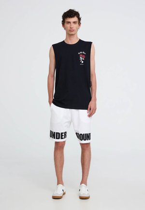 Canotta nera con design di rose, abbinata a pantaloni corti bianchi con testo nero "UNDERGROUND". Calzature: sneakers bianche con accenti marroni.