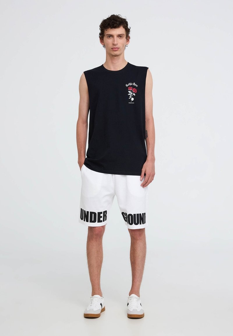 Canotta nera con design di rose, abbinata a pantaloni corti bianchi con testo nero "UNDERGROUND". Calzature: sneakers bianche con accenti marroni.