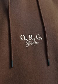 Haut à capuche marron avec une texture douce, cordons de serrage aux embouts noirs et logo brodé "O.R.G. Studio" en blanc.
