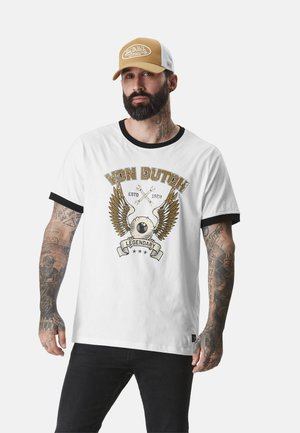 T-shirt en coton blanc avec finition noire, arborant un design graphique en or et noir de ailes et le texte "VON DUTCH LEGENDARY". Porté avec une casquette beige et blanche.