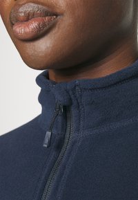 Marine fleece pullover met een hoge kraag en ritssluiting. Kenmerken zijn flatlock naden en een getextureerde stof. Close-up van de nek en lippen.