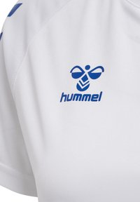 Sigla albină albastră Hummel și text pe material alb de îmbrăcăminte sport, cu cusături vizibile și o parte a mânecii.