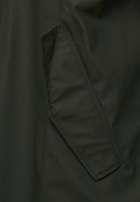 Gros plan d'une poche en tissu vert foncé avec rabat et coutures visibles sur un vêtement d'extérieur.