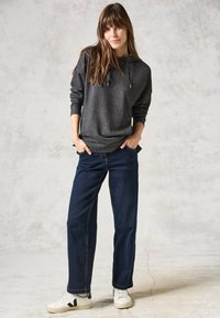 Grijze hoodie met trekkoorden, gecombineerd met donkerblauwe jeans. Model draagt witte sneakers met zwarte accenten. Eenvoudige en casual outfit.