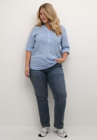 Blusa azul claro de manga larga con cuello en forma de pico y tapeta de botones, con un patrón de lunares. Combinada con jeans azul oscuro y zapatillas deportivas.