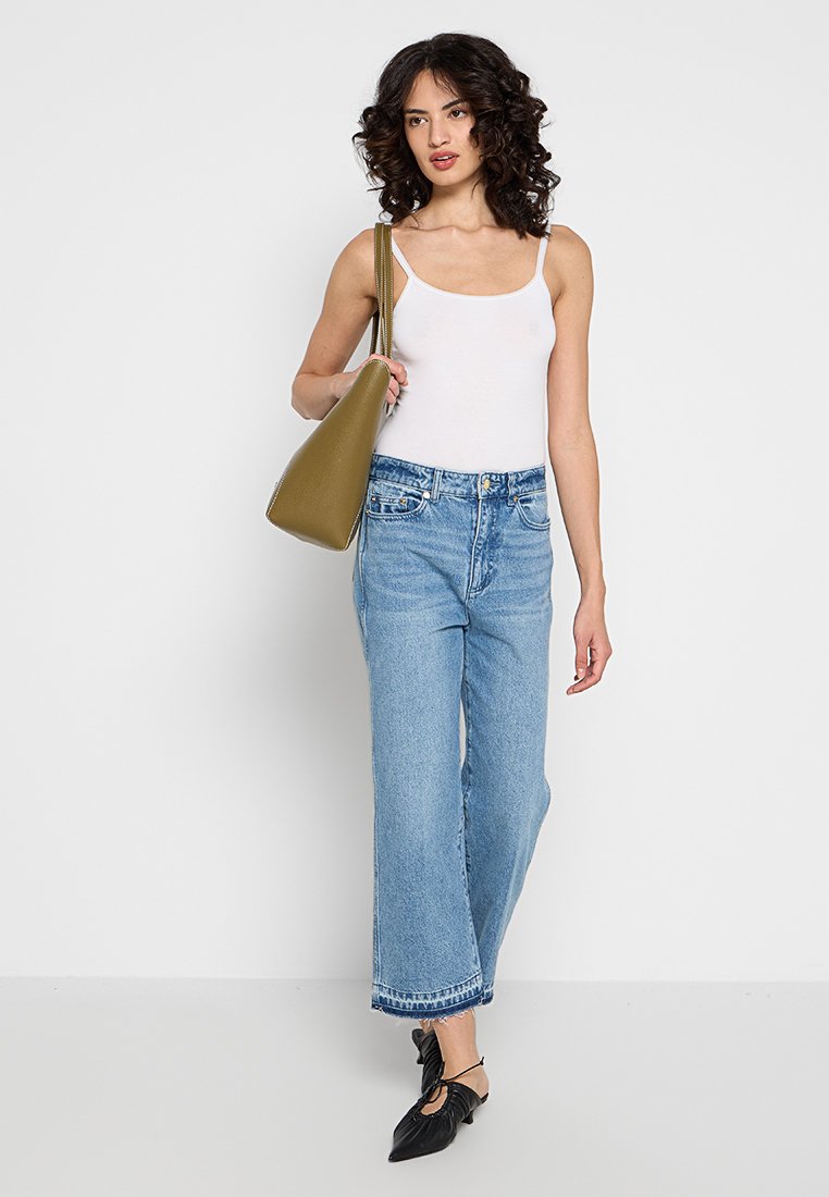 Top ajustado blanco, jeans de mezclilla de pierna ancha azules, zapatillas planas negras con detalle de nudo y un bolso verde olivo. Texturas suaves, estilo casual.