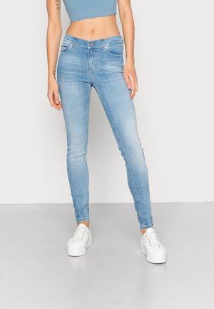 Kvinde iført lyseblå skinny jeans, hvide platform-sneakers og en kort blå tanktop, stående mod en ensartet lys baggrund.