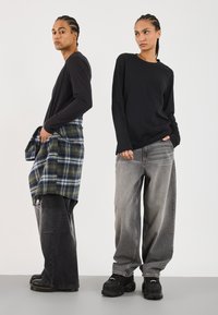 Zwei Personen tragen langärmlige schwarze Shirts. Eine hat ein kariertes grün-blaues Hemd um die Taille gebunden; beide tragen weite graue Jeans und schwarze Schuhe.