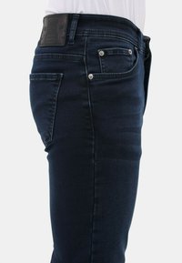 Donkerblauwe nauwsluitende denim jeans met voor- en achterzakken, zilveren klinknagels en een zwarte leren patch met schedelontwerpen op de tailleband.