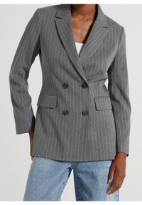 Femme portant un blazer gris à rayures fines croisé avec poches à rabat, par-dessus un haut blanc et un jean bleu clair, sur un fond uni.