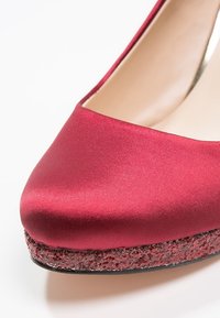 Chaussure à talon haut en satin rouge avec bout arrondi et semelle plate-forme rouge pailletée, vue en gros plan sur fond blanc.