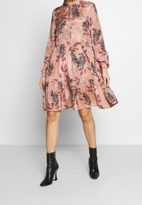 Robe rose à manches longues avec un ourlet à volants, ornée de motifs de feuilles noires. Le tissu semble soyeux avec un léger éclat. Une paire de bottes à talons noires.