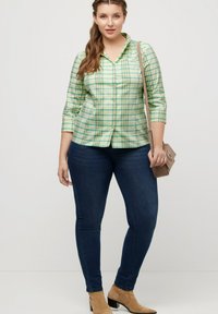 Camisa de cuadros verde y azul de manga tres cuartos, combinada con jeans skinny oscuros y botas de tobillo marrones; sosteniendo un bolso pequeño.