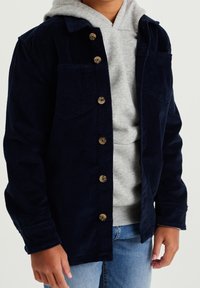 Námořnická kordová bunda s knoflíkovým zapínáním vpředu, dvěma náprsními kapsami a límečkem, nošená přes šedou mikinu a světle modré džíny.