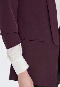 Tumši violeta žakete ar gludu tekstūru, ar stilīgu griezumu, sānu kabatu un baltu, pūkainu, garām piedurknēm internetu blūzi zem tā.