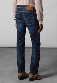 Jeans in denim blu scuro con una vestibilità dritta, dotati di tasche posteriori e di un'etichetta in pelle marrone sulla vita. Stivali in pelle con texture a pelle di serpente visibili.