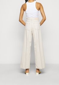 Beige wide-leg trousers made of cotton, featuring a high waist and back pockets. Paired with a white sleeveless top and light-colored heels.