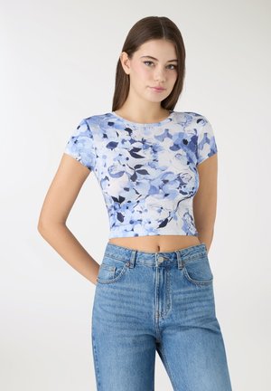 Krótoptop w kwiatowy wzór na jasnoniebieskim tle z ciemnoniebieskimi i czarnymi kwiatami. Modelka zestawiła go z wysokim stanem jeansów.