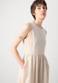 Beige sleeveless dress with a round neckline and gathered waistline. Features side pockets, made of a smooth, lightweight fabric.