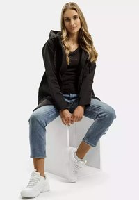 Czarna wodoodporna kurtka z kapturem, noszona na czarnej bluzce, w połączeniu z jasnoniebieskimi, skróconymi dżinsami i białymi sneakersami. Model siedzi na pudełku.