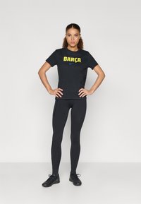 Μαύρη μπλούζα με κίτρινο λογότυπο "BARÇA" και σήμα της Nike, συνδυασμένη με μαύρα κολάν και μαύρα αθλητικά παπούτσια. Απλός, εφαρμοστός σχεδιασμός.