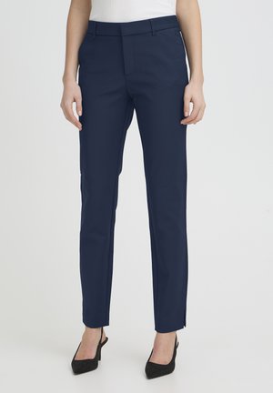 Femme portant un pantalon bleu marine ajusté et des talons noirs à bout pointu, debout devant un fond clair uni.