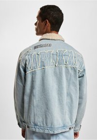 Světle modrá džínová bunda s kožichem z umělé kožešiny, s opotřebením a vyšitými nápisy "KARL KANI" a "EST.1989" na zadní straně.