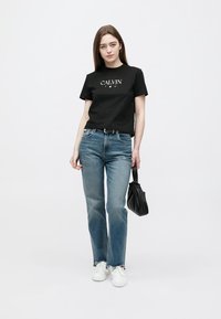 Čierne bavlnené tričko s nápisom "CALVIN", modré džínsy z denimu a biele tenisky. Modelka drží čiernu kabelku; minimalistický dizajn, stredne priliehavý strih.