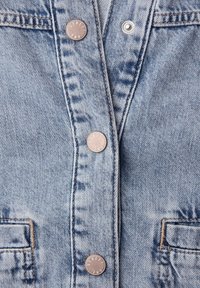 Detailní záběr světle modré džínové bundy s kovovými patentními knoflíky a viditelným šitím kolem knoflíkové klopy a kapes.