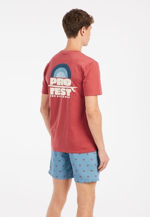 Rode katoenen T-shirt met een grafisch ontwerp op de achterkant, met de tekst "PRO TEST" en een boog. Gecombineerd met blauwe shorts met een palmbomenpatroon.