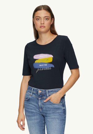 Femme portant un t-shirt noir avec des coups de pinceau pastel et le texte "Gardez la vision, faites confiance au processus," associé à un jean bleu.