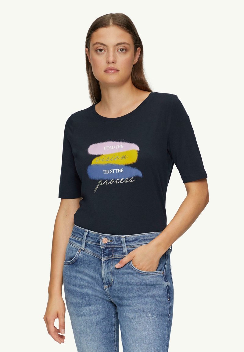 Vrouw draagt een zwart t-shirt met pastel penseelstreken en de tekst "Houd de visie vast, vertrouw op het proces," gecombineerd met een blauwe spijkerbroek.