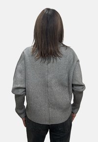 Una persona con capelli castani di media lunghezza indossa un maglione grigio a maglia e jeans scuri, vista di spalle su uno sfondo semplice.