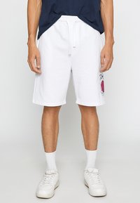 Pantalones cortos de algodón blancos con cintura elástica y cordón. Tiene bolsillos laterales y un gráfico rojo en el muslo izquierdo. Se lleva con zapatillas blancas.