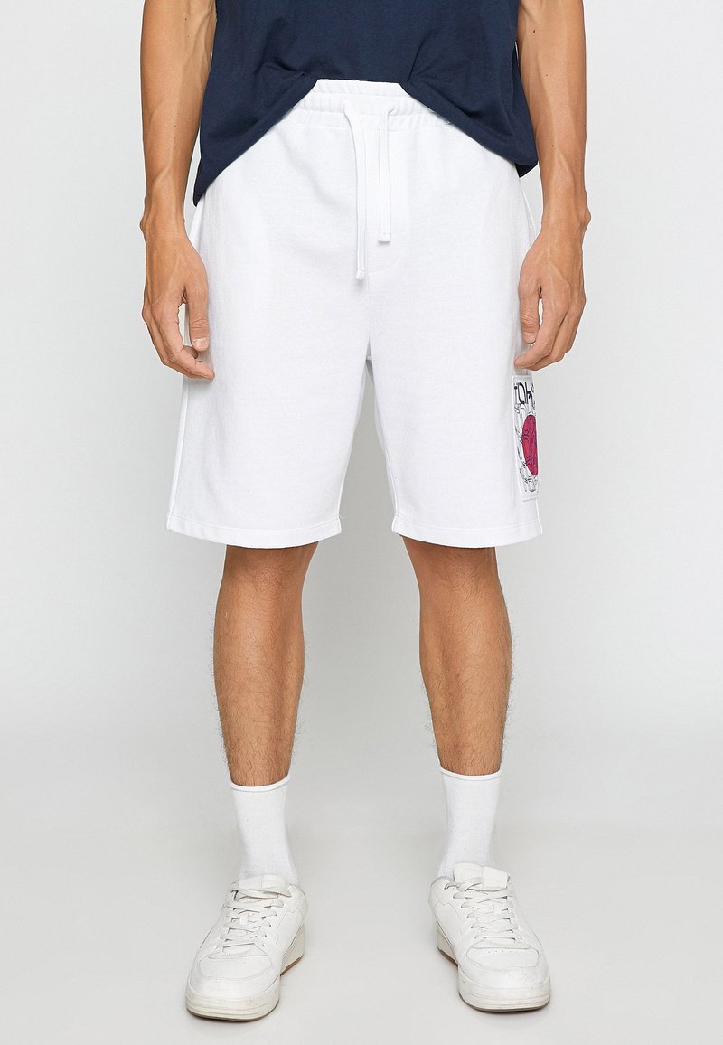 Pantalones cortos de algodón blancos con cintura elástica y cordón. Tiene bolsillos laterales y un gráfico rojo en el muslo izquierdo. Se lleva con zapatillas blancas.