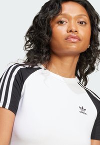 Adidas balta marškinėliai su juodais raglanų rankovėmis, turi tris baltas juostas kiekvienoje rankovėje ir mažą juodą logotipą ant krūtinės.