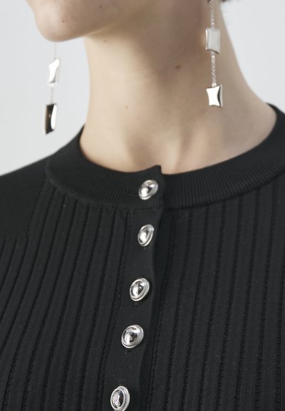 Zwarte geribbelde top met een ronde kraag en zilveren decoratieve knopen. Lange zilveren hangers met geometrische accenten zijn zichtbaar.