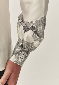 Chemise blanche avec des détails floraux noirs complexes en dentelle sur le poignet. Tissu lisse, coupe ajustée et texture froncée au niveau du poignet.