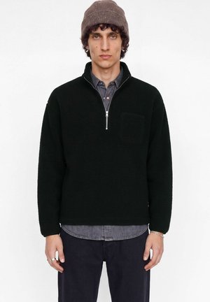 Un homme pose sur fond blanc, vêtu d’un pull noir à demi-zip par-dessus une chemise grise, d’un pantalon foncé et d’un bonnet en maille marron.