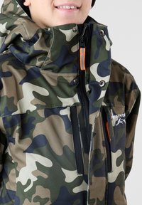 Camo-gepatroneerde jas in groen, bruin en zwart met een hoge kraag, ritssluiting en een oranje accent op de rits. Bevat een logopatch.