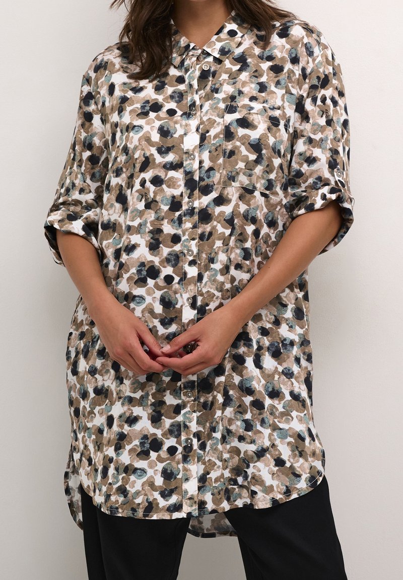 Femme portant une chemise ample à col et boutons, manches retroussées, avec un motif abstrait multicolore tacheté en beige, noir et bleu sarcelle.