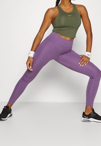 Žena v olivově zeleném tílku bez rukávů, fialových legínách, bílých náramcích a černých teniskách Nike, která si široce protahuje nohy na jednobarevném pozadí.