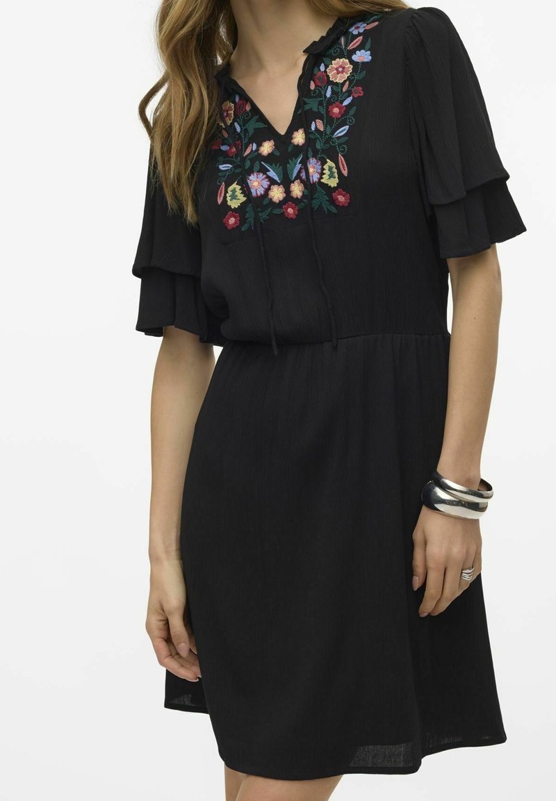 Svart klänning med korta, volangärmar; har färgglad blomstermönstring runt halsringningen; lätt och flödande tyg.