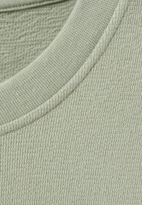 Gros plan sur un tissu vert clair en maille gaufrée avec un col côtelé, montrant des coutures texturées et détaillées.