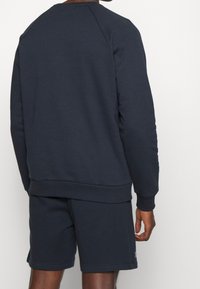 Uomo che indossa una felpa blu navy scuro e pantaloni corti coordinati, visto da dietro su uno sfondo bianco semplice.